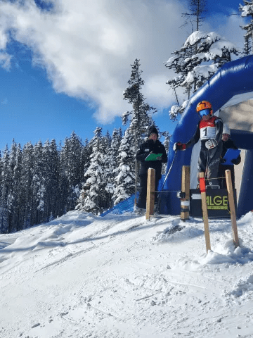 A skier stands at the start of a slope, ready to race, with a timing gate in the background. Snow-covered trees and a blue sky create a scenic backdrop.