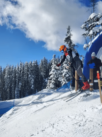 Skier in bright gear launching off a snow ramp under a blue sky, surrounded by evergreen trees, showcasing winter sports action.