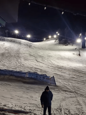 A snowy ski slope illuminated at night, with a lone figure standing in the foreground. Ski tracks are visible, indicating activity on the hill.